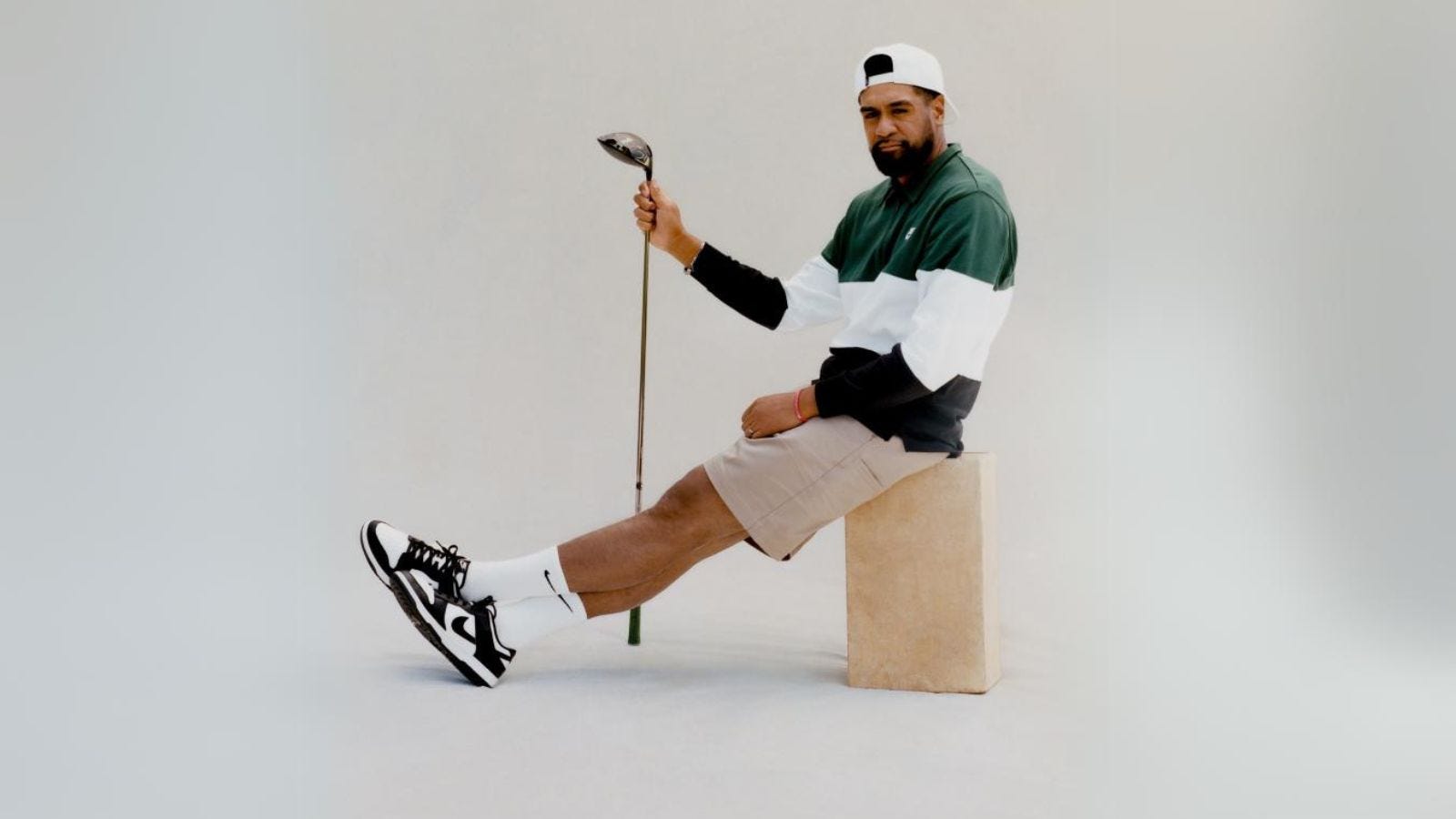 Tony Finau sitting down, holding a golf club and wearing a striped polo shirt, tan shorts, and Nike sneakers